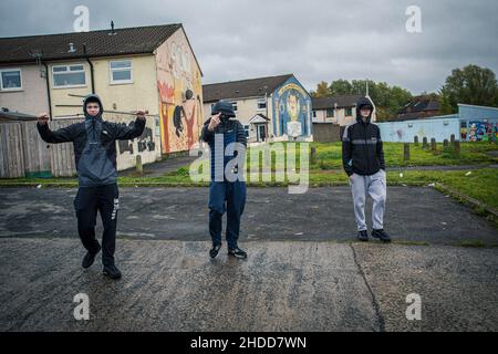 Des jeunes marchant devant des peintures murales politiques avec des symboles paramilitaires sur le Lower Shankill Estate, à Belfast. Banque D'Images