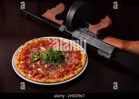 Un pistolet à sous-machine Thompson sur une table brun foncé et une pizza au jambon avec du fromage Banque D'Images