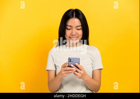 Positive adorable fille brune asiatique à cheveux longs dans un t-shirt décontracté, utilise son smartphone, messagerie avec des amis en ligne, parcourt les réseaux sociaux, sourit, se tient sur un arrière-plan orange isolé Banque D'Images