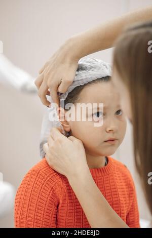 gros plan des mains d'esthéticienne mettant blanc capuchon médical jetable sur la tête de six ans-fille dans le bureau de cosmétologie, se préparant pour le perçage d'oreilles Banque D'Images