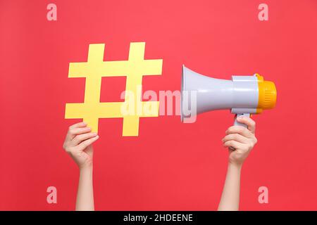 Gros plan des mains de femmes tenant le signe jaune hashtag et le mégaphone, étiquette pour les affaires, le marketing et la publicité, isolé sur fond rouge studio.Con Banque D'Images