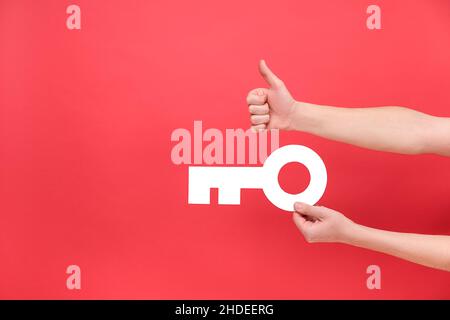 Gros plan de mains de sexe masculin tenant une grande clé blanche et montrant le pouce vers le haut, excellent symbole, isolé sur un mur d'arrière-plan rouge dans un studio avec espace de copie Banque D'Images