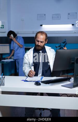 Medic tenant la radiographie pour examen tout en prenant des notes sur le document de contrôle.Médecin rédigeant le diagnostic et les résultats après l'analyse par radiographie pour un traitement médical à minuit. Banque D'Images