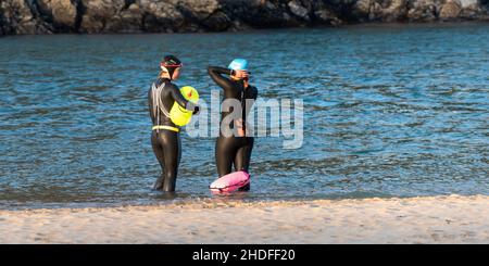 Une image panoramique de deux nageurs sauvages femelles portant des combinaisons et utilisant des flotteurs de remorquage gonflables en fluorescent dans la rivière Gannel sur Crantock Beach, au nord-est Banque D'Images