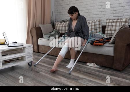 Jeune femme pensive assise sur le canapé regardant sa jambe, après une chirurgie de l'ACL. Banque D'Images