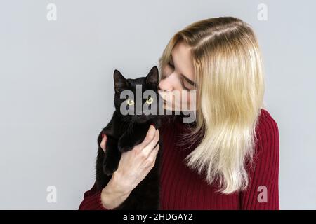 Jeune caucasien joli blond mignon en rouge tenant le chat noir, comme le bébé, isolé sur fond clair.La fille garde le chaton.Amitié entre l'animal et le propriétaire Banque D'Images