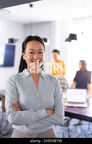 Portrait d'une femme d'affaires asiatique souriante aux bras croisés tandis que ses collègues discutent en arrière-plan Banque D'Images