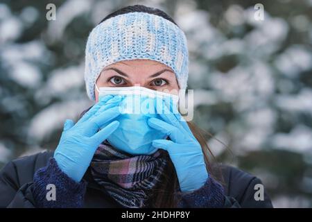 Jeune femme en hiver blouson avec bleu jetable à usage unique masque de virus du visage et gants de protection, neige floue couvert arbres fond, gros plan det Banque D'Images