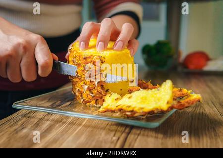 Gros plan d'une jeune femme coupant un ananas juteux avec un couteau de chef sur une table en bois dans une cuisine. Banque D'Images