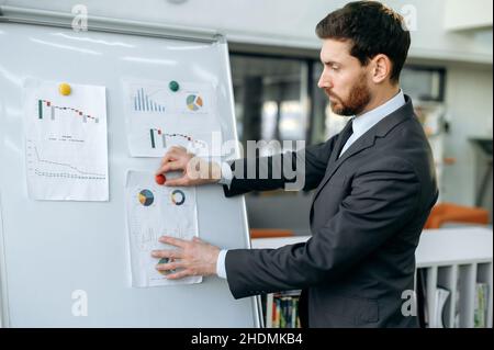 Pensive intelligent homme d'affaires caucasien adulte, analyste financier, chef de département, se tient près du tableau blanc dans le bureau, analyse des tableaux financiers, réfléchit les risques financiers, les bénéfices, la stratégie d'entreprise Banque D'Images