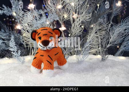 Un tigre tricoté en peluche se dresse sur la neige artificielle et des arbres bleus et blancs artificiels avec des lumières festives se tiennent derrière eux sur un fond bleu foncé Banque D'Images