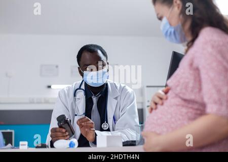 Praticien qui fait la consultation avec la patiente enceinte, portant des masques pour la protection.Femme qui attend un enfant et qui reçoit des conseils médicaux sur la grossesse de la part d'un médecin pendant une pandémie Banque D'Images