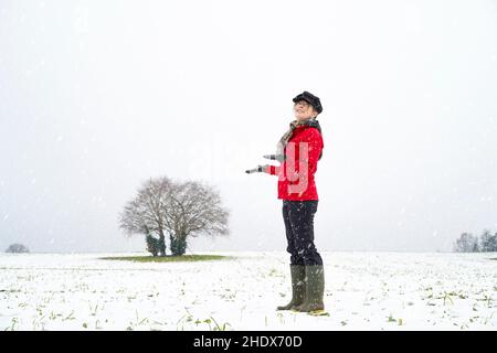 La jeune femme souriante, avec sa veste rouge vif et son chapeau tendance, se tient isolée dans les champs de campagne, se tenant les mains à l'extérieur et regardant la neige qui tombe en hiver. Banque D'Images