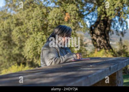 femme aux cheveux blancs avec des lunettes prenant des notes dans son carnet dans le parc lors d'un beau jour d'hiver Banque D'Images