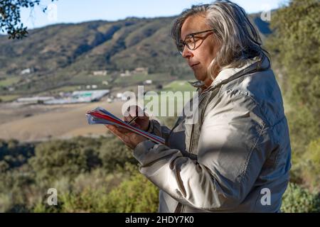 femme aux cheveux blancs avec des lunettes prenant des notes dans son carnet dans le parc lors d'un beau jour d'hiver Banque D'Images