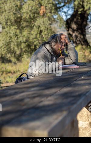 femme aux cheveux blancs avec des lunettes prenant des notes dans son carnet dans le parc lors d'un beau jour d'hiver Banque D'Images