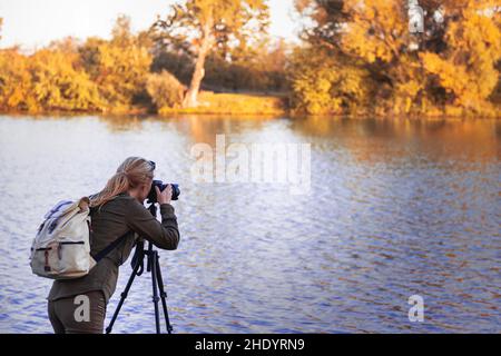 La femme prend des photos de paysage à l'automne.Randonneur avec appareil photo sur trépied à côté du lac. Banque D'Images