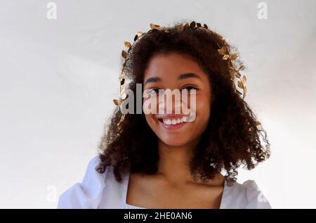 Gros plan portrait d'une femme afro-américaine mignonne avec des cheveux bouclés dans un serre-tête à feuilles d'or, avec un aspect flirté à l'appareil photo isolé sur blanc, jolie femme avec Banque D'Images