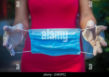 Démonstration du masque médical bleu clair.Mains en gants blancs.Silhouette féminine en robe rouge hors du foyer en arrière-plan.Masque médical et gants blancs Banque D'Images