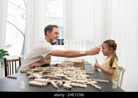 Vue latérale du papa vérifiant la température corporelle avec thermomètre infrarouge frontal de la fille assise à la table et jouant avec des blocs de bois Banque D'Images