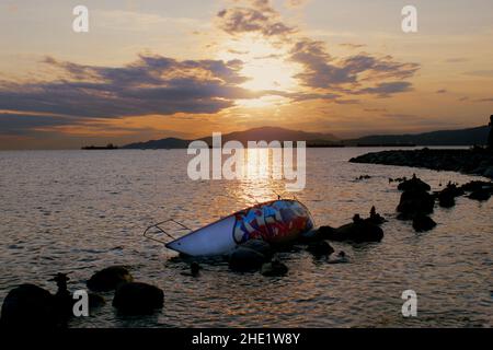 Un bateau couvert de graffitis a chaviré au coucher du soleil dans une baie calme, avec des montagnes et des navires ancrés silhouettes sur un ciel nocturne spectaculaire. Banque D'Images