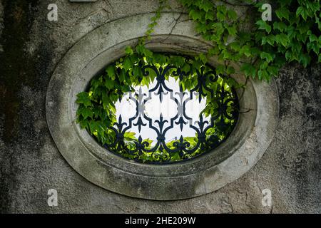 Une fenêtre ovale avec une grille de sécurité artisanale dans un mur, surcultivée par le vin sauvage. Banque D'Images