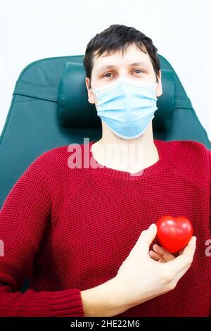 Le jeune homme en masque médical s'assoit dans la chaise et tient le petit coeur dans sa main.Patient reconnaissant.Sujet des soins de santé et du don.Sauvetage des malades Banque D'Images