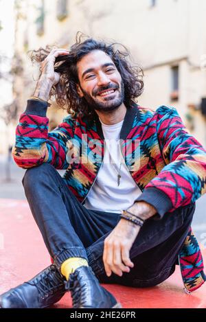 Homme hispanique barbu dans des vêtements élégants et décontractés touchant les cheveux maussants. En regardant l'appareil photo avec le sourire, assis à pieds croisés sur le banc de la rue de la ville Banque D'Images