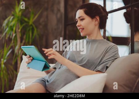 Jeune femme enceinte souriante tenant une table numérique et effectuant un appel vidéo tout en étant assise sur le lit à la maison, parlant en ligne avec sa famille ou ses amis.Distance p Banque D'Images