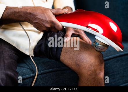 Gros plan d'un vieil homme méconnaissable utilisant un appareil de massage de vibration pour la douleur articulaire du genou à la maison - concept de machine de soulagement de la douleur et thérapie ou Banque D'Images