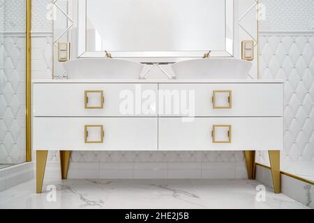 Armoire blanche avec poignées et jambes dorées et lavabos élégants avec grand miroir décoré avec motif losange dans la salle de bains de près Banque D'Images