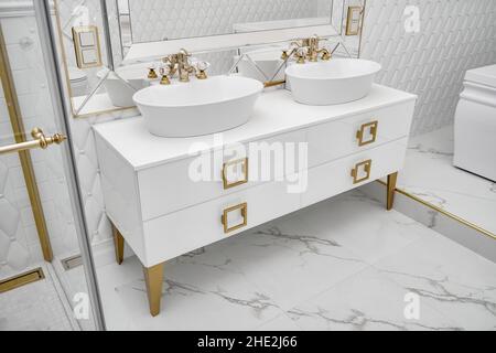 Armoire blanche avec poignées et jambes dorées et lavabos élégants avec grand miroir décoré avec motif losange dans la salle de bains de près Banque D'Images