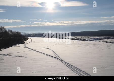 Paysage enneigé ensoleillé en hiver.Le soleil brille dans le ciel bleu au-dessus de la rivière gelée et enneigée.Pistes pour véhicules tout-terrain sur une vaste surface enneigée. Banque D'Images