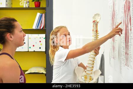 Jeune femme patiente à la consultation de physiothérapie, physiothérapeute expliquant les os du dos sur le schéma du corps des muscles humains, tenant le modèle de colonne vertébrale Banque D'Images