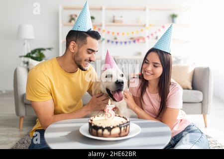 Joyeux et divers conjoints célébrant l'anniversaire de leur chien avec un gâteau savoureux, portant des chapeaux de fête à la maison.Fête des fêtes Banque D'Images