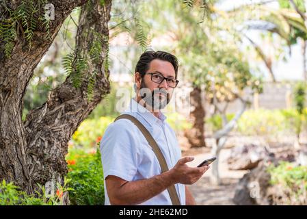 Portrait d'un homme barbu adulte utilisant un téléphone portable dans le parc portant des vêtements professionnels et des lunettes regardant sur l'appareil photo - des gens de race blanche Banque D'Images