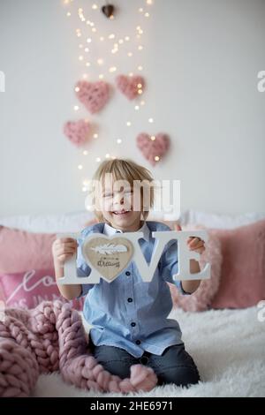 Mignon enfant blond avec chemise, assis sur le lit avec décoration de Saint-Valentin, arrière-plan isolé Banque D'Images