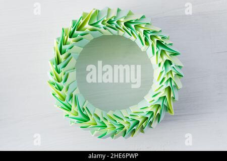 Anneau d'origami, structure circulaire paramétrique abstraite composée de feuilles de papier vertes et jaunes, pose sur un bureau blanc, vue du dessus Banque D'Images