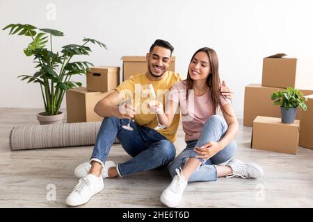 Couple multiracial millénaire ayant une fête d'armement ménager, assis sur le sol avec du champagne, célébrant l'achat de la nouvelle maison Banque D'Images