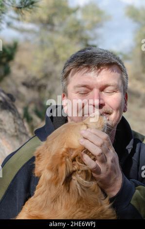 Portrait d'un chien rouge mixte et d'un homme mature.L'animal de compagnie est assis dans les bras de son propriétaire et léche ses lèvres.Homme d'âge moyen avec cheveux gris courts et stu Banque D'Images