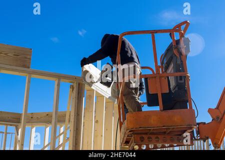 Gros plan d'un travailleur dans des gants de protection blancs tenant une planche l'installant sur la construction de ménage à l'aide d'un marteau pneumatique dans des poutres en bois clouant. Banque D'Images