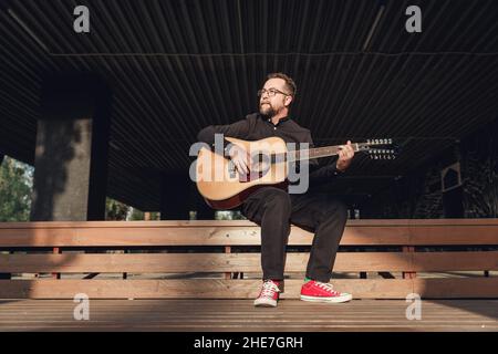 Homme en noir avec une barbe jouant de la guitare acoustique à 12 cordes sur un terrain urbain en bois en plein air en été. Banque D'Images