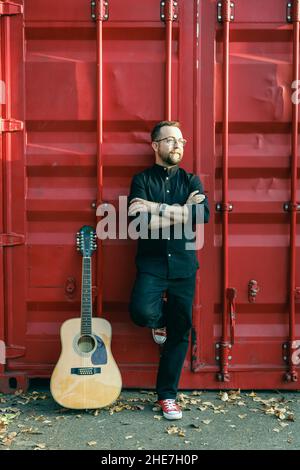 Homme en noir avec une barbe penchée contre le récipient rouge avec une guitare acoustique à 12 cordes Banque D'Images