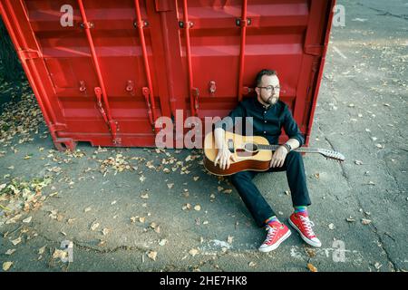 Homme en noir avec une barbe assise sur un sol sous un récipient rouge avec guitare acoustique à 12 cordes Banque D'Images