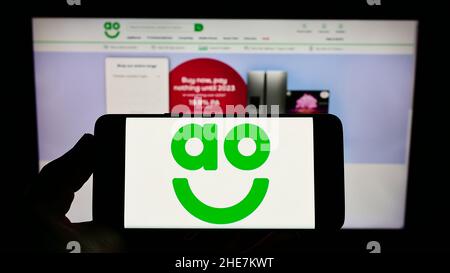 Personne tenant un téléphone portable avec le logo de la société britannique de vente au détail en ligne AO World plc à l'écran en face de la page Web d'affaires.Mise au point sur l'affichage du téléphone. Banque D'Images