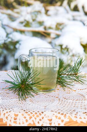 Infusion de thé à aiguille de pin chaud dans une tasse de thé en verre transparent sur la table.Pins enneigés en arrière-plan, en plein air par temps froid d'hiver. Banque D'Images