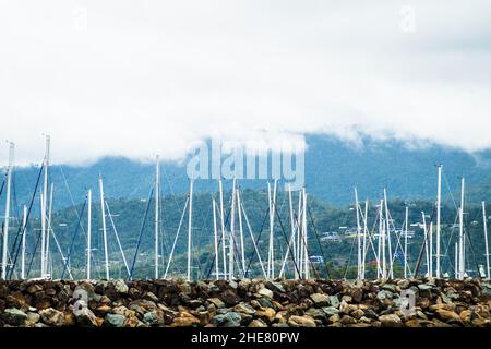 Mâts de yachts amarrés dans la marina protégée par un mur de rochers et avec un fond de montagne à nuages bas parsemé de maisons, Airlie Beach, Queensland Banque D'Images