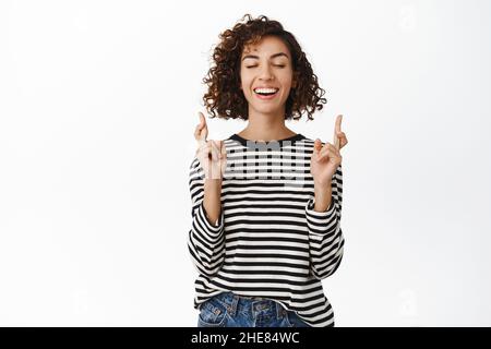 Image d'une fille heureuse pleine d'espoir souhaitant, priant, croisant les doigts pour la bonne chance, aspirant à réaliser quelque chose, se tenant sur fond blanc Banque D'Images