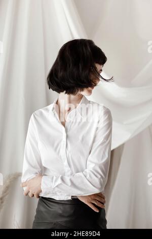 Sympathique et confiante jeune femme aux bras pliés souriant à l'appareil photo et s'amusant. Fille en chemise blanche et jupe en cuir assis à la maison studio. Banque D'Images