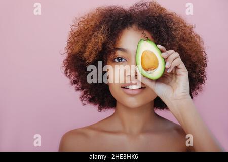 Portrait d'une belle jeune femme à la peau sombre avec des cheveux bouclés couvre un oeil avec la moitié d'un avocat regarde l'appareil photo et sourit sur un fond rose. Banque D'Images